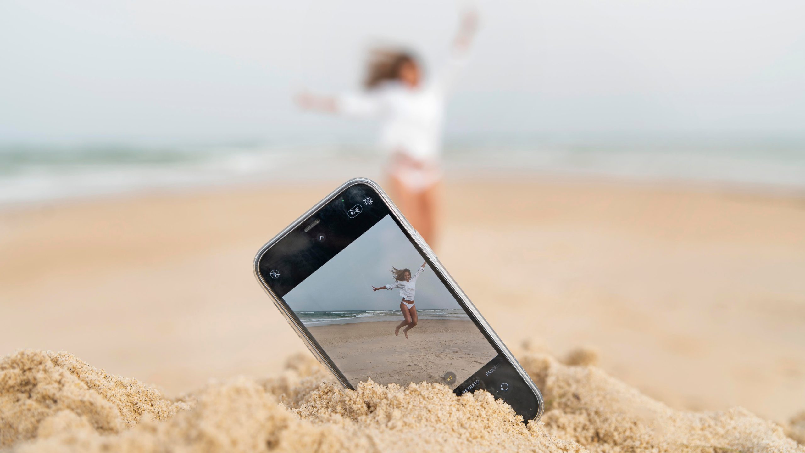 Una mujer saltando en la playa mientras su movil toma la foto enterrado en la arena.
